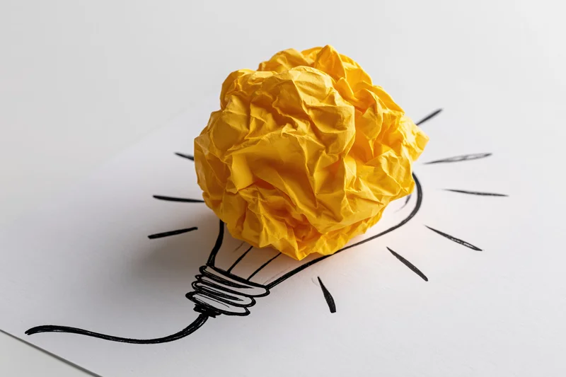 Crumpled yellow paper ball placed on hand-drawn lightbulb sketch on white paper