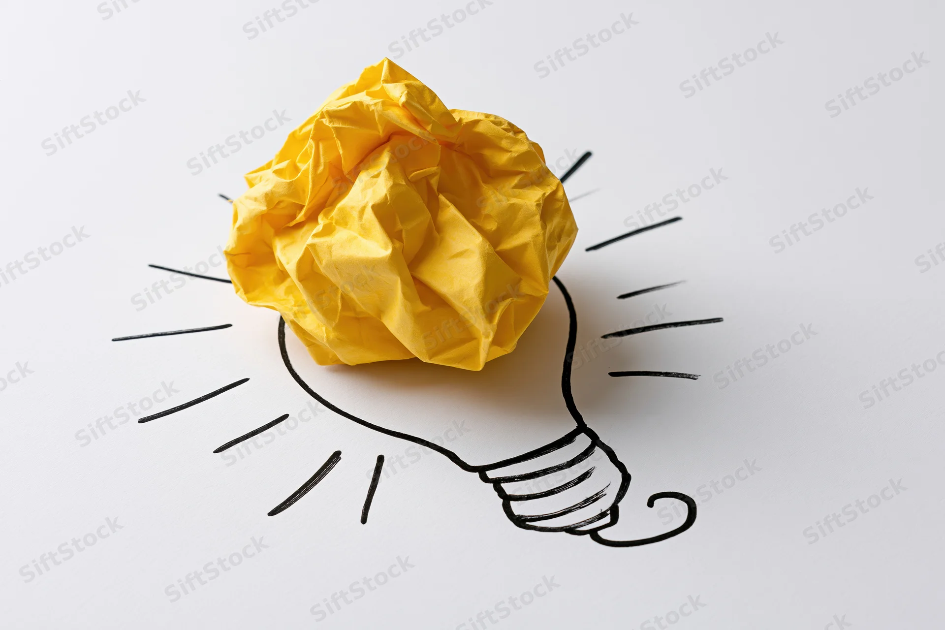 Crumpled yellow paper ball placed on a hand-drawn lightbulb sketch on white surface