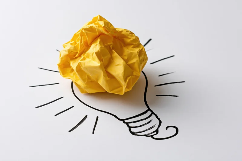 Crumpled yellow paper ball placed on a hand-drawn lightbulb sketch on white surface