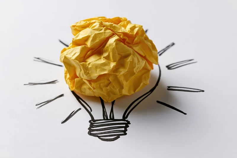 Crumpled yellow paper ball placed on hand-drawn lightbulb sketch on white surface