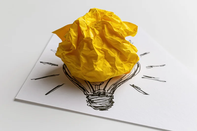 Crumpled yellow paper ball resting on hand-drawn lightbulb sketch on white paper