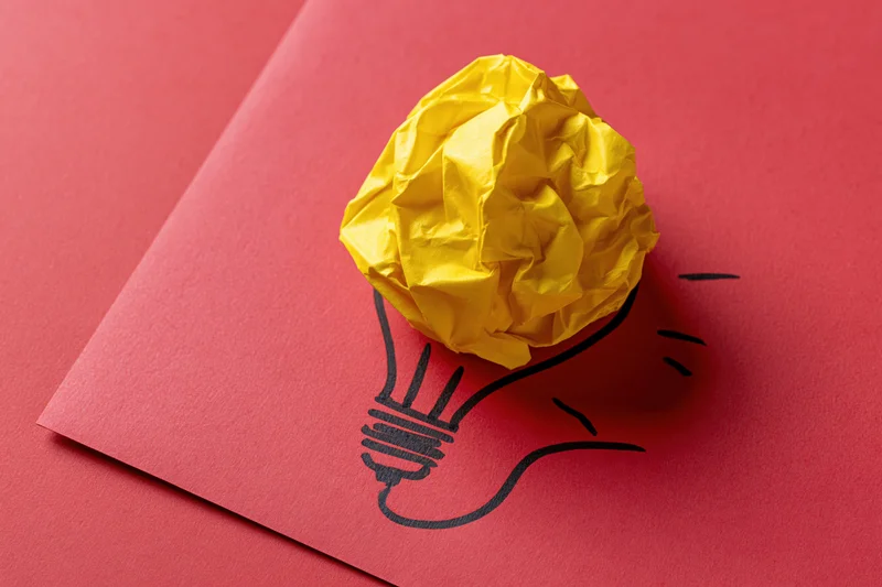 Crumpled yellow paper ball placed on red cardboard with hand-drawn lightbulb outline