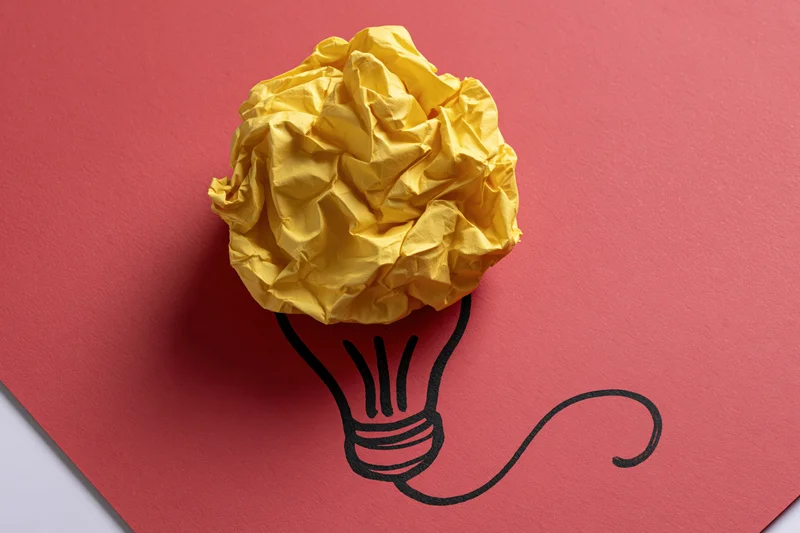 Crumpled yellow paper ball placed on red cardboard with hand-drawn lightbulb outline