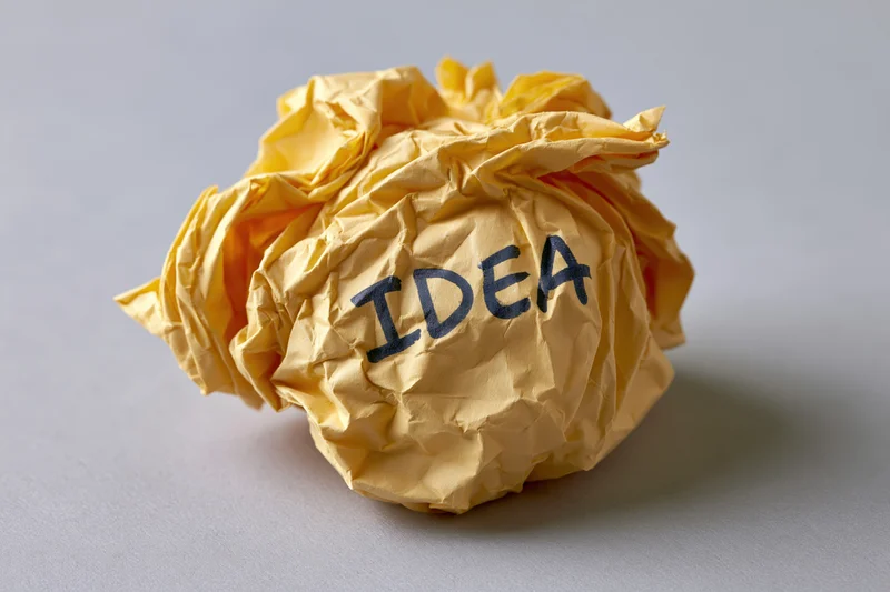 Crumpled yellow paper ball with handwritten idea text on gray surface