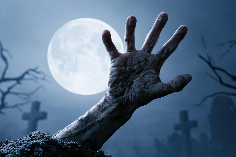 Decayed zombie hand emerging from soil in a graveyard under a full moon at night