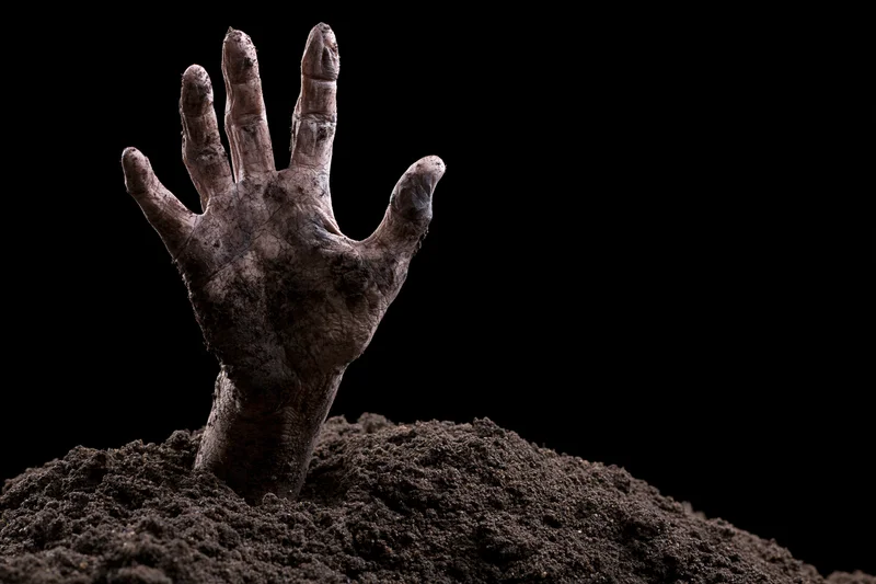 Decayed hand clawing upward from a mound of dirt against black background