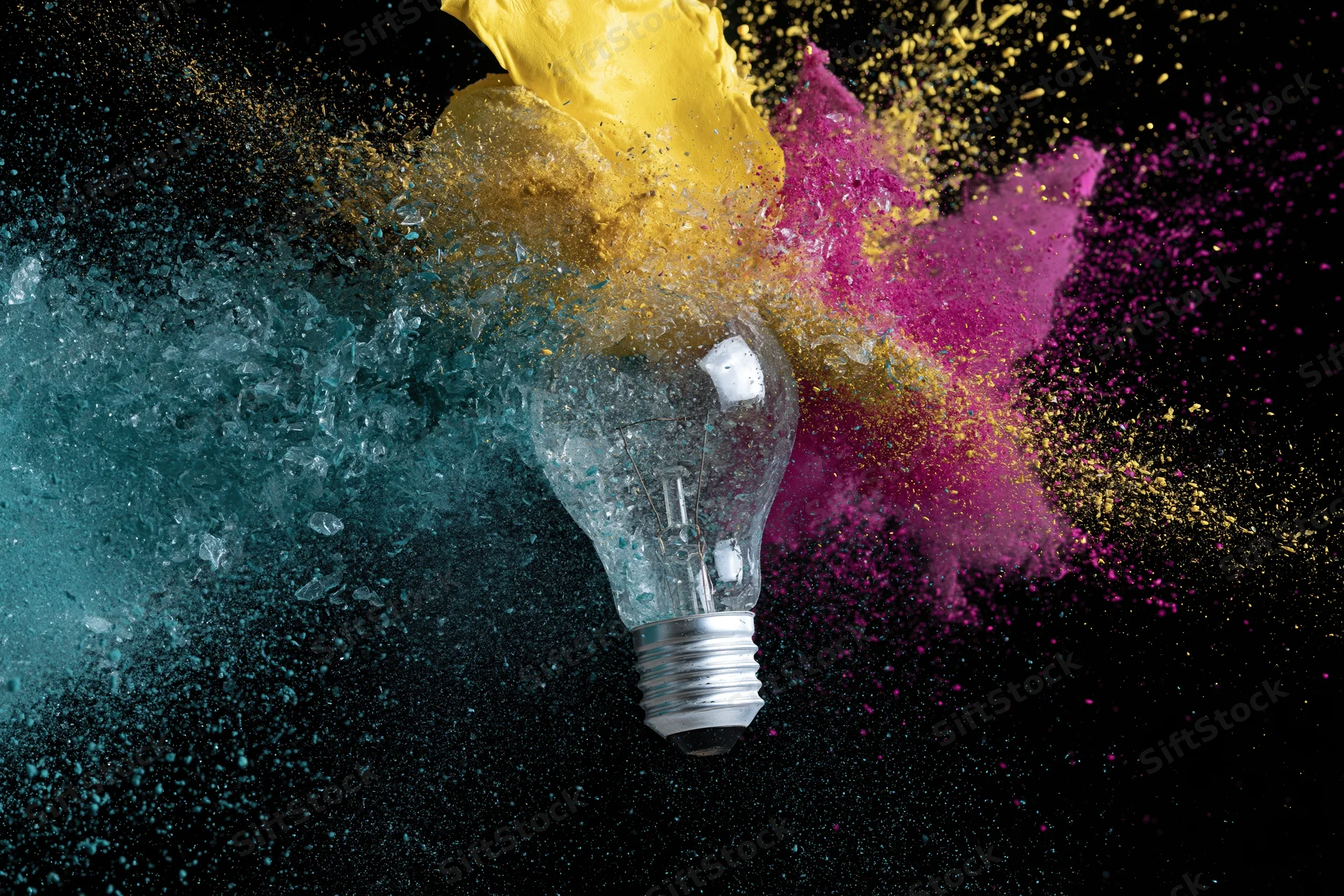Lightbulb shattering mid-air with colorful powder exploding outward against black background