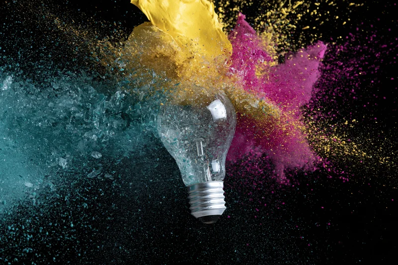 Lightbulb shattering mid-air with colorful powder exploding outward against black background