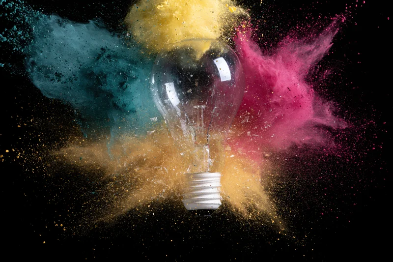 Glass lightbulb exploding mid-air with colorful powder burst against black background