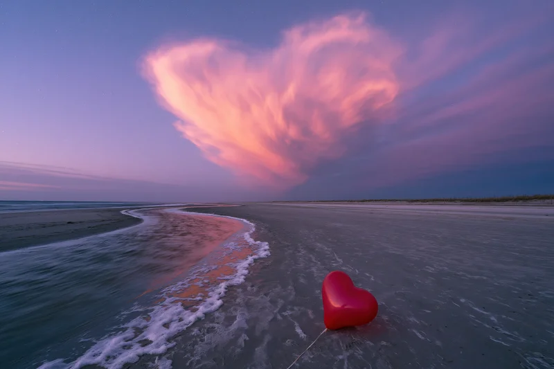 Red heart-shaped balloon resting on sandy beach at twilight with pink clouds above