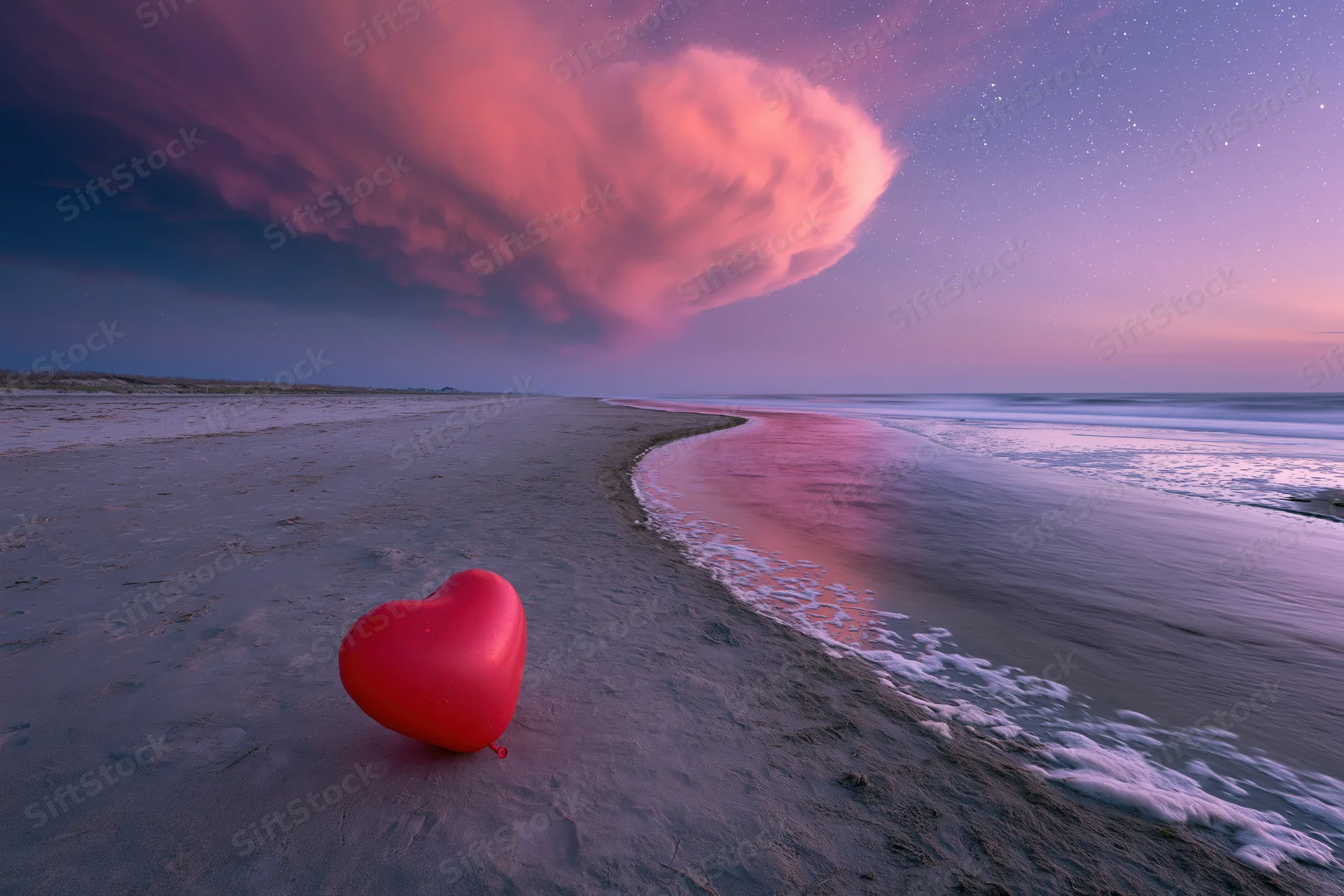 Red heart-shaped balloon resting on sandy beach beside ocean waves under starry twilight sky