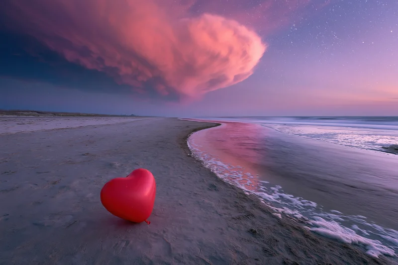Red heart-shaped balloon resting on sandy beach beside ocean waves under starry twilight sky