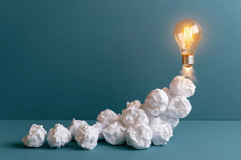 Glowing lightbulb rising from a pile of crumpled paper on a blue surface