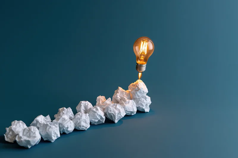 Glowing lightbulb rising from a row of crumpled paper balls on a blue surface