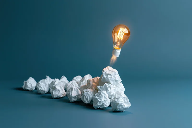 Glowing lightbulb launching upward from pile of crumpled paper on blue background