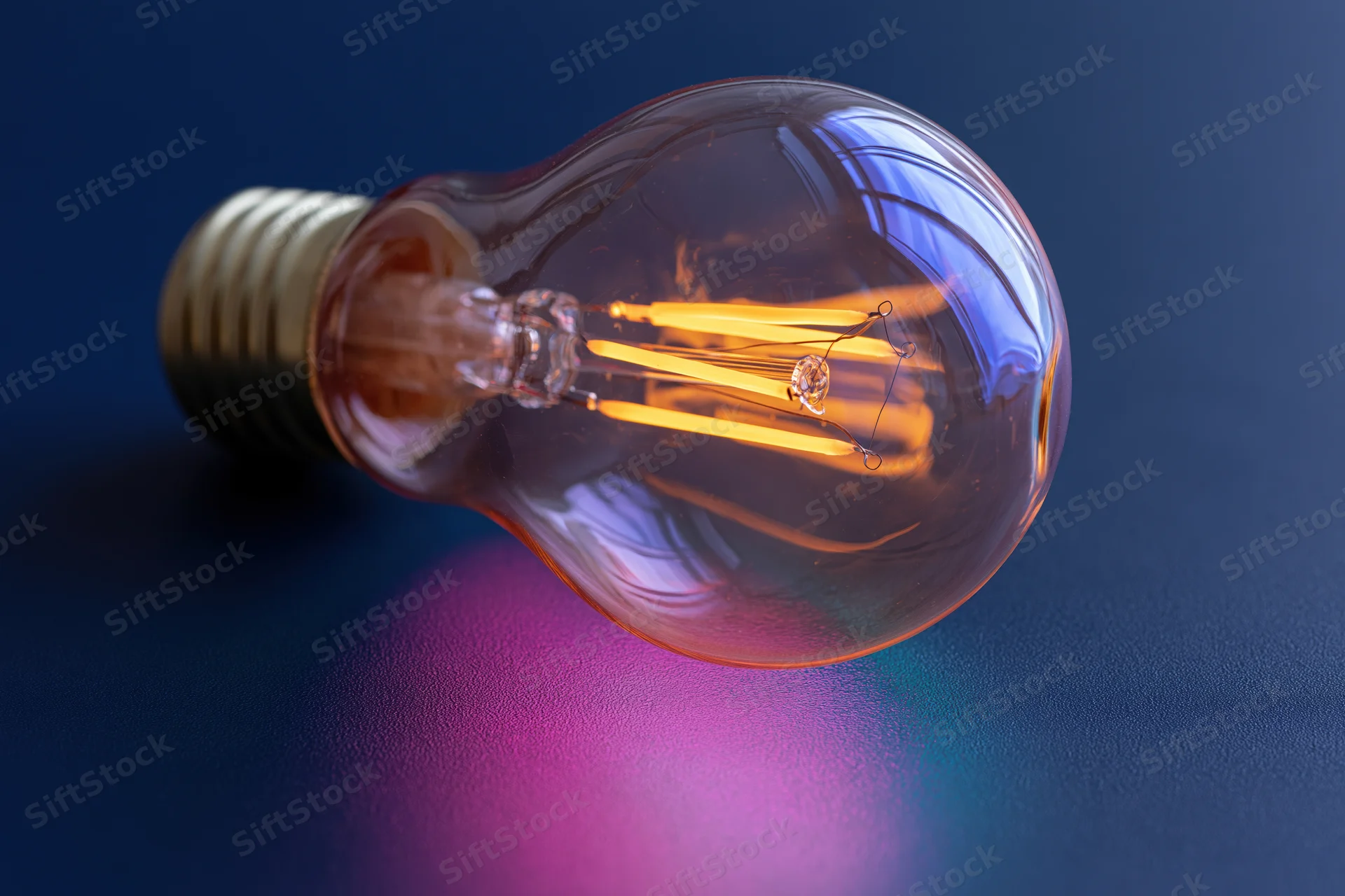 Glowing incandescent light bulb lying on its side on a dark surface with colorful reflections