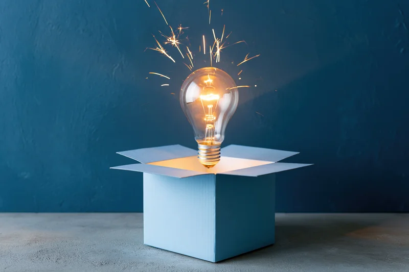Glowing lightbulb emerging from open blue box with sparks against dark blue wall