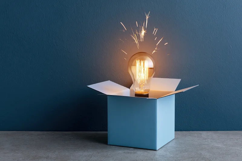 Glowing lightbulb emerging from open blue box on gray floor against dark blue wall
