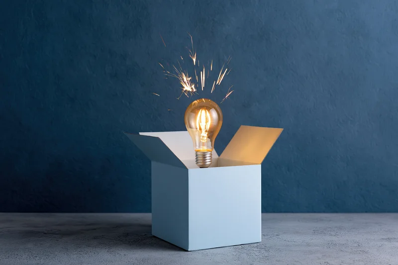 Glowing incandescent light bulb with sparks rising from open box on concrete surface against blue wall