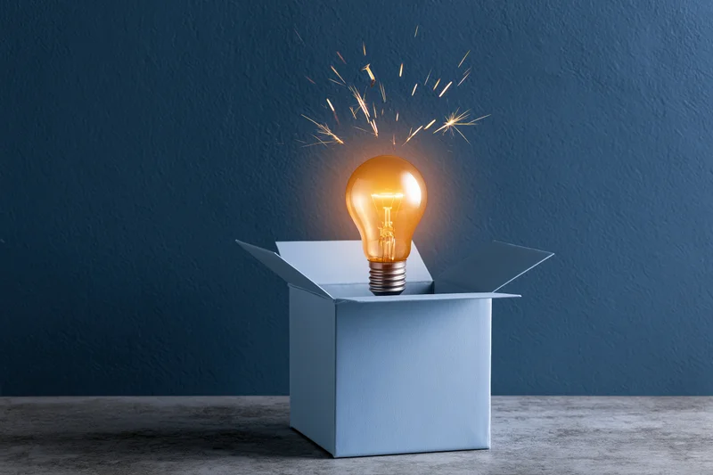 Glowing lightbulb emerging from open box on dark blue wall