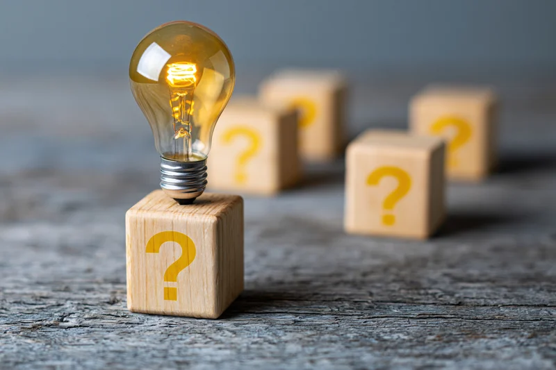 Glowing lightbulb on wooden cube with question mark, surrounded by similar cubes on textured surface