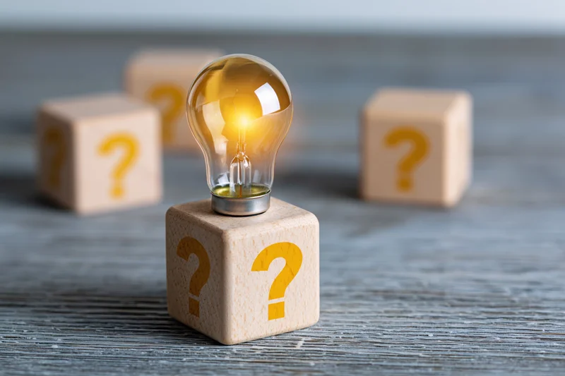 Glowing incandescent light bulb on wooden cube with question marks, surrounded by similar cubes on textured gray surface