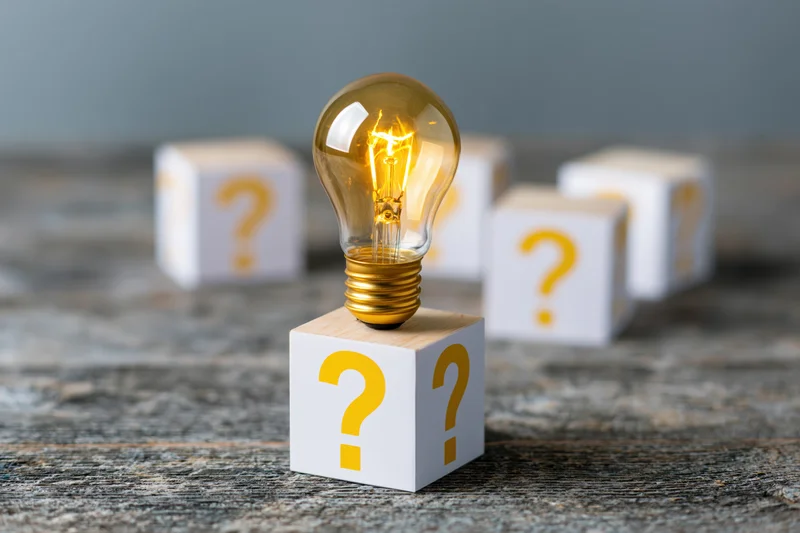 Glowing incandescent light bulb on a wooden cube with question marks, surrounded by similar cubes on a textured surface
