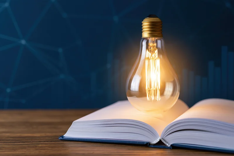Glowing incandescent lightbulb resting on open book on wooden surface