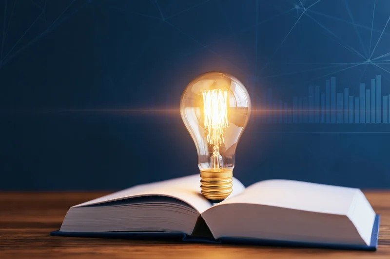 Glowing incandescent lightbulb resting on open book on wooden table with digital network and data graph in background