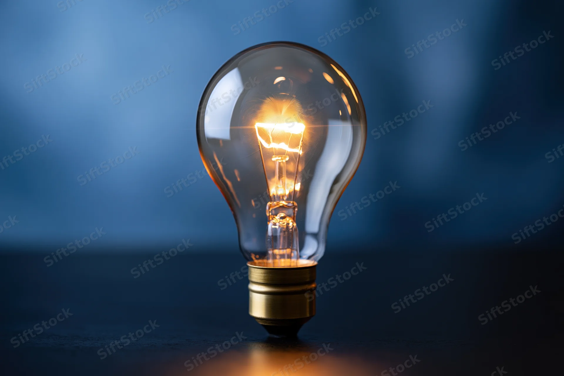 Glowing incandescent light bulb on dark reflective surface with blue background