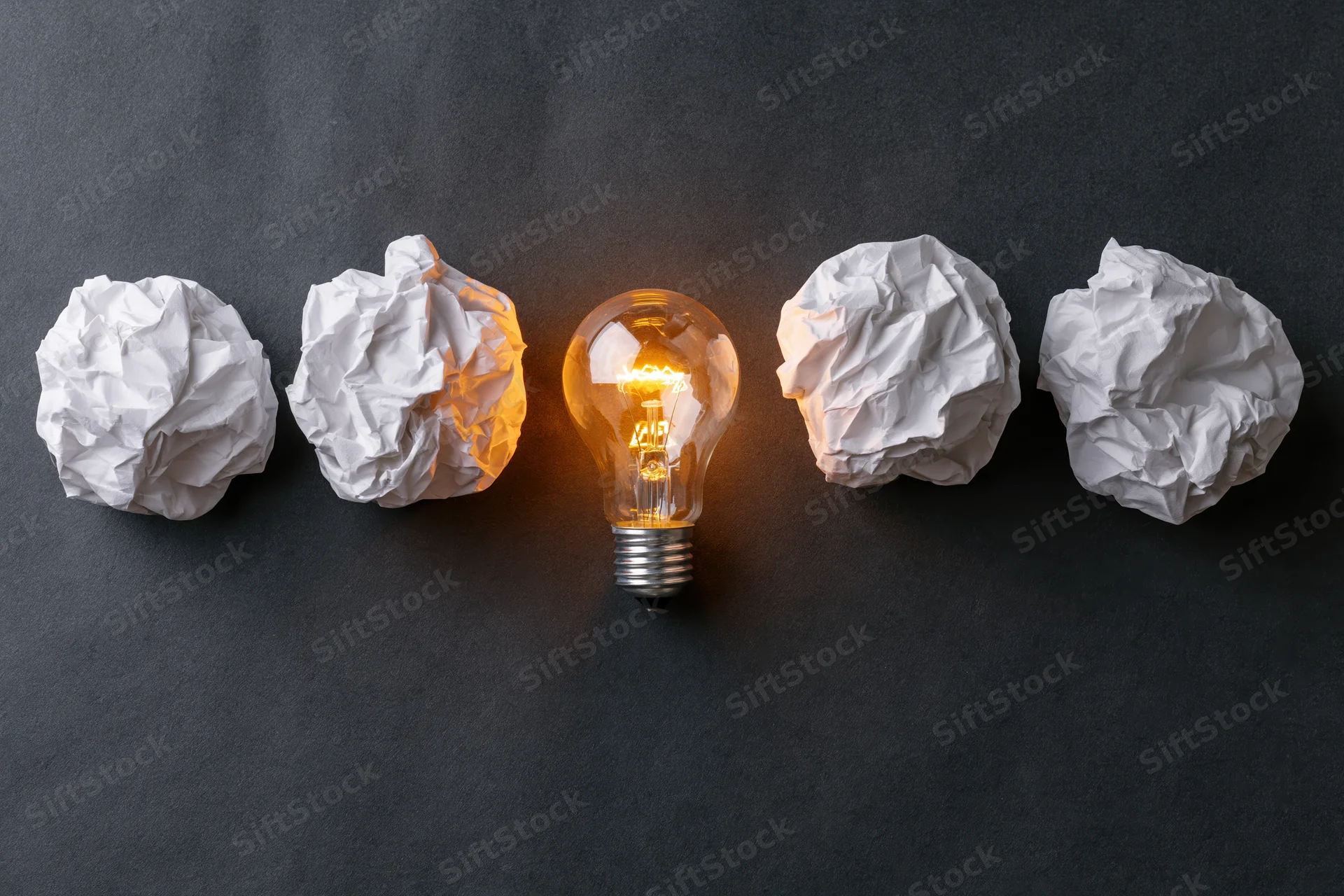 Glowing lightbulb centered among crumpled paper balls on dark surface
