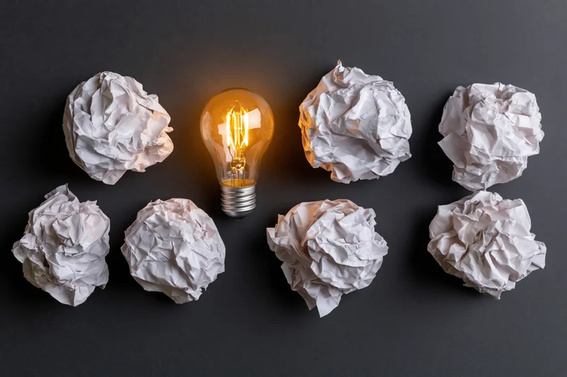 Glowing lightbulb surrounded by crumpled paper balls on dark surface