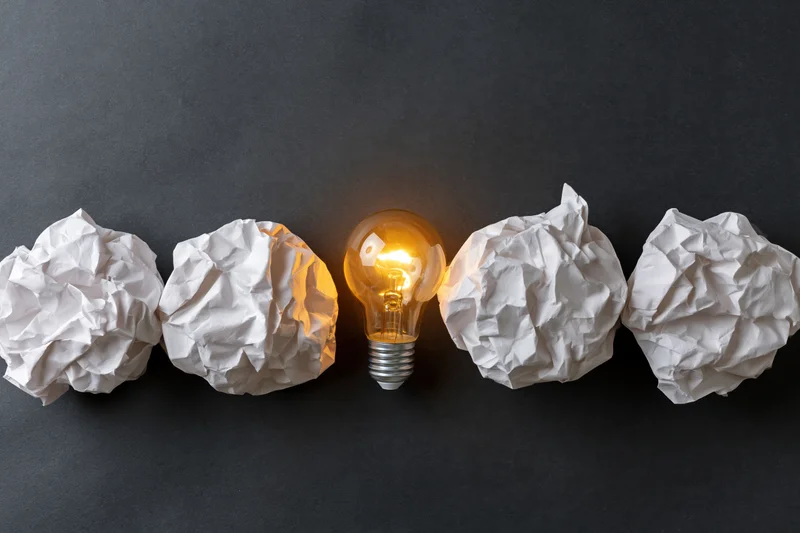 Glowing lightbulb among crumpled paper balls on dark surface
