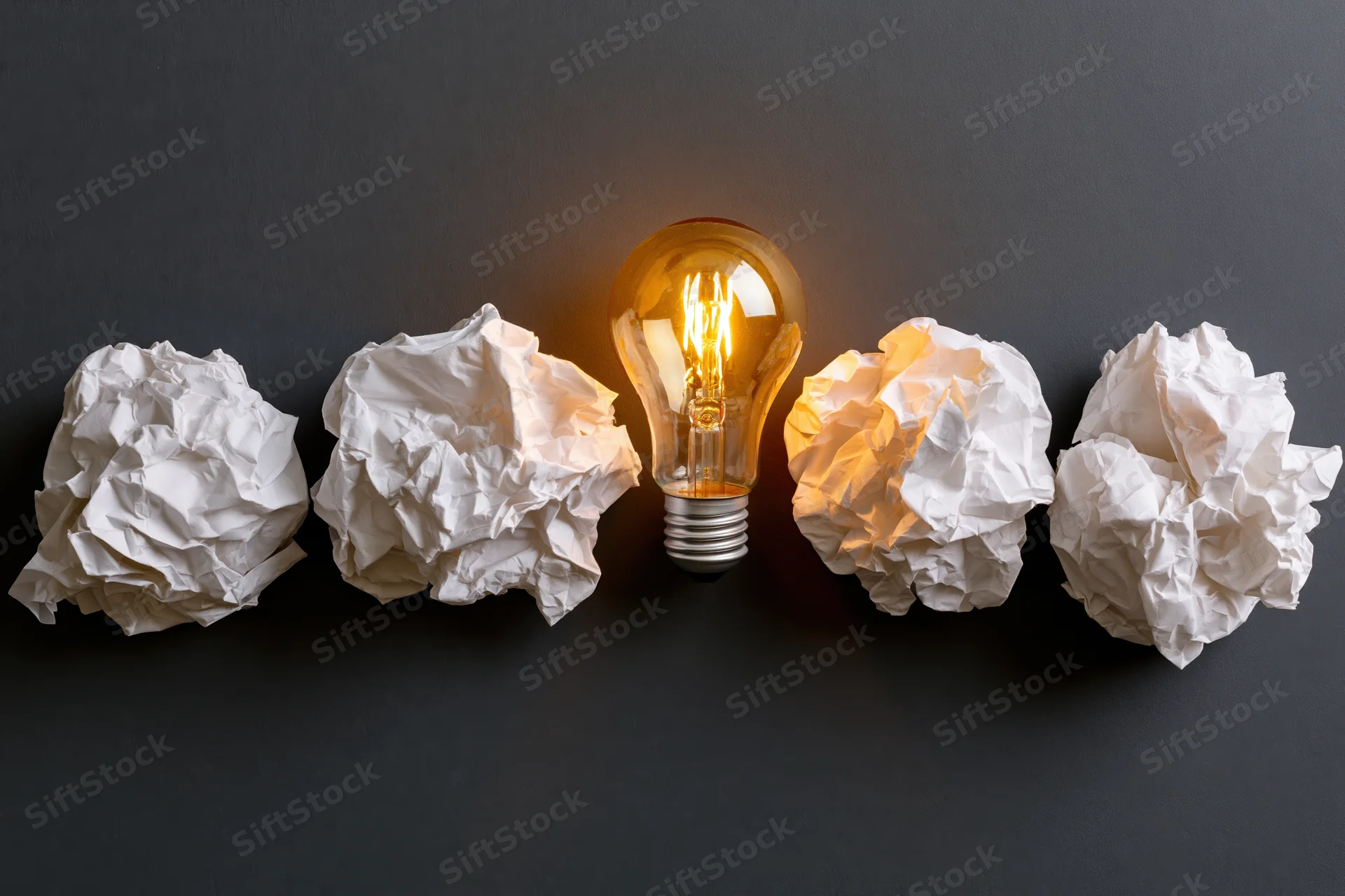 Glowing lightbulb centered among crumpled paper balls on dark surface