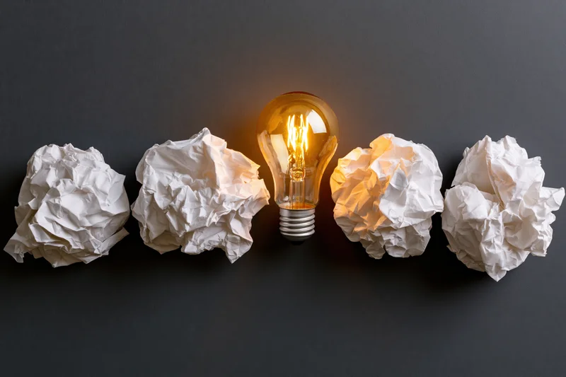 Glowing lightbulb centered among crumpled paper balls on dark surface