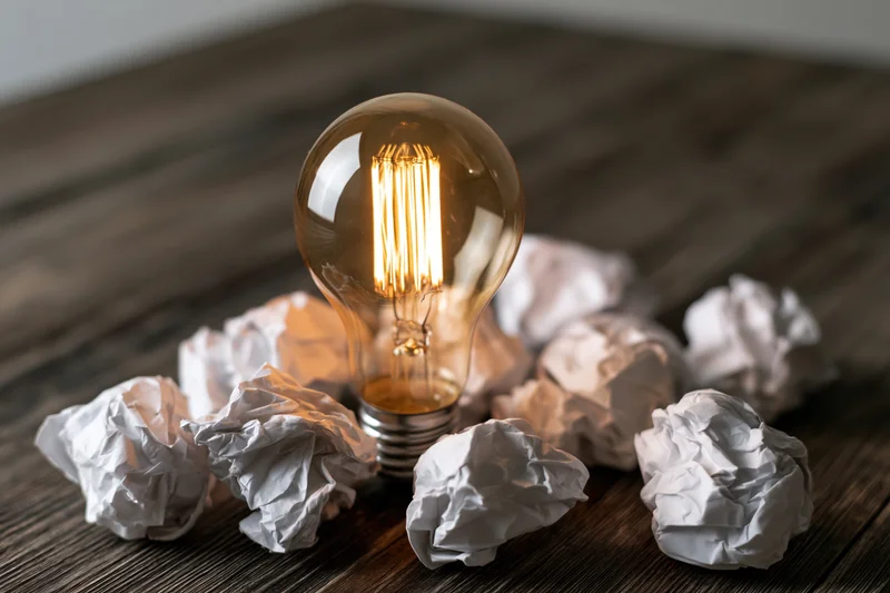 Glowing incandescent lightbulb surrounded by crumpled paper on a dark wooden table