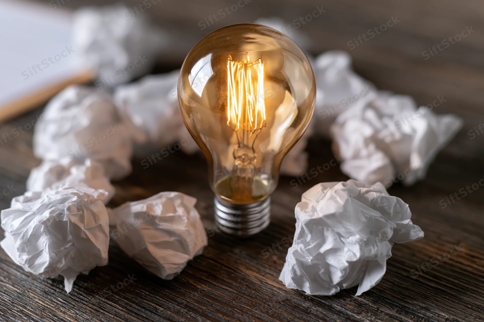 Glowing incandescent lightbulb surrounded by crumpled paper on a dark wooden desk