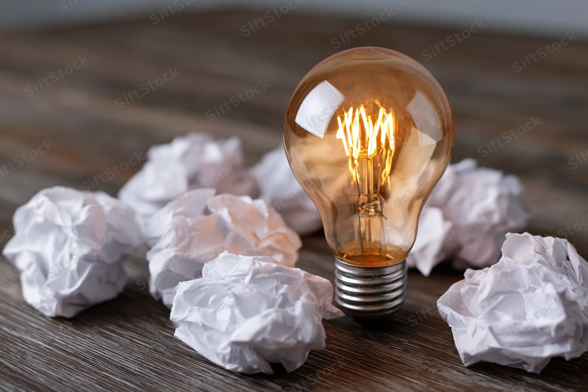 Glowing incandescent lightbulb surrounded by crumpled paper on wooden desk