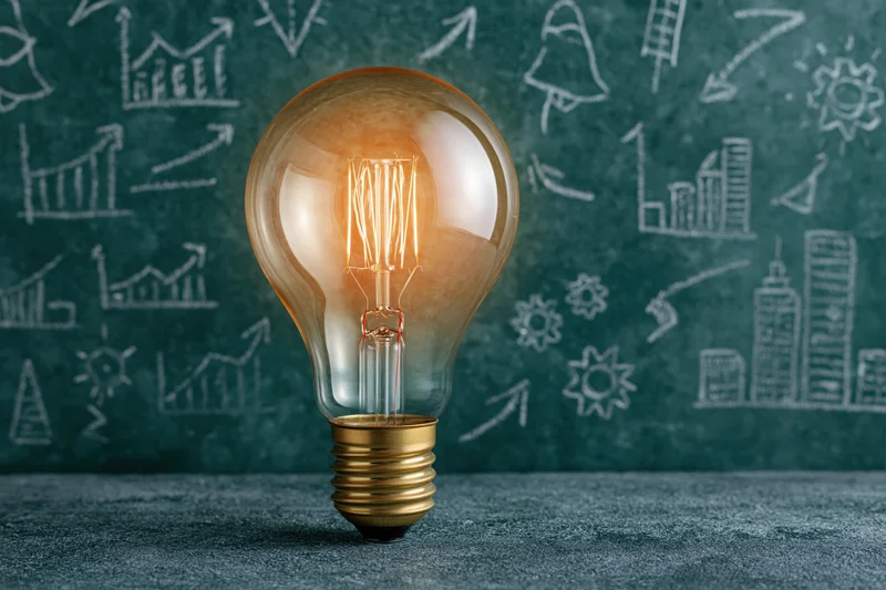 Glowing incandescent lightbulb with brass base on dark surface in front of chalkboard with business and innovation drawings