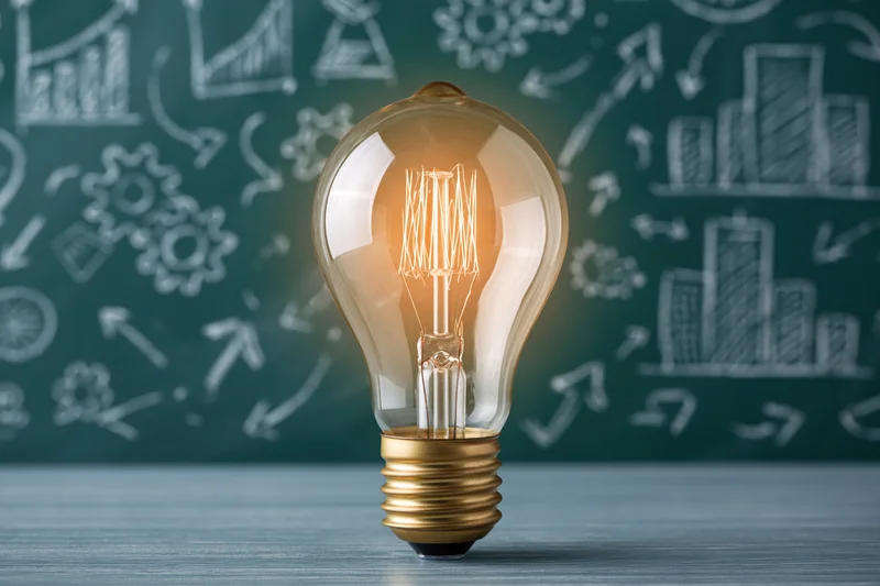 Glowing incandescent lightbulb with brass base on wooden desk in front of chalkboard with business sketches