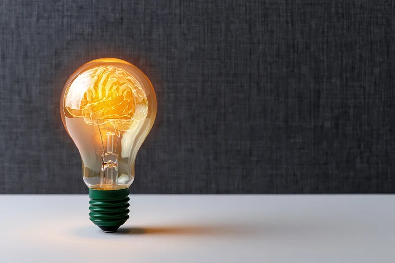 Glowing lightbulb with brain shape inside on white surface against gray background