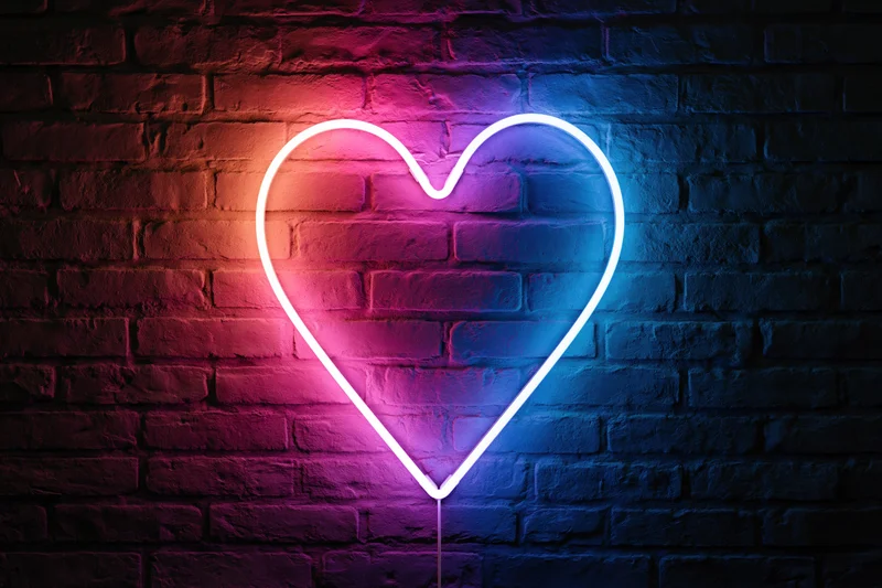 Neon heart sign glowing in pink and blue gradient light on dark brick wall