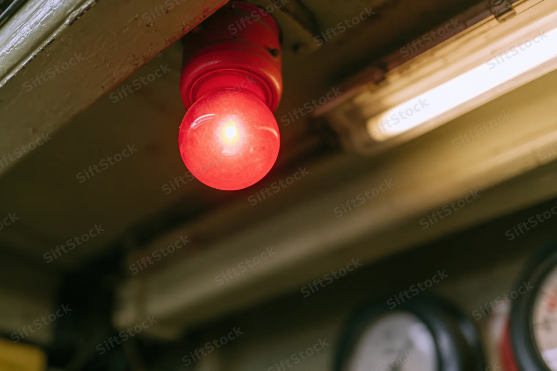 Glowing red signal bulb mounted beneath metal industrial ceiling with gauges and fluorescent light