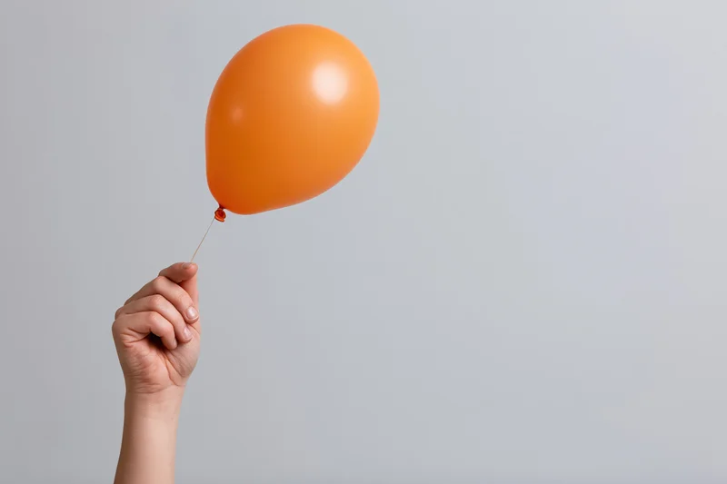 Hand holding an orange balloon by its string against a plain gray background