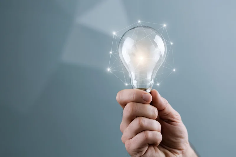 Hand holding a glowing lightbulb with digital network overlay against a gray background