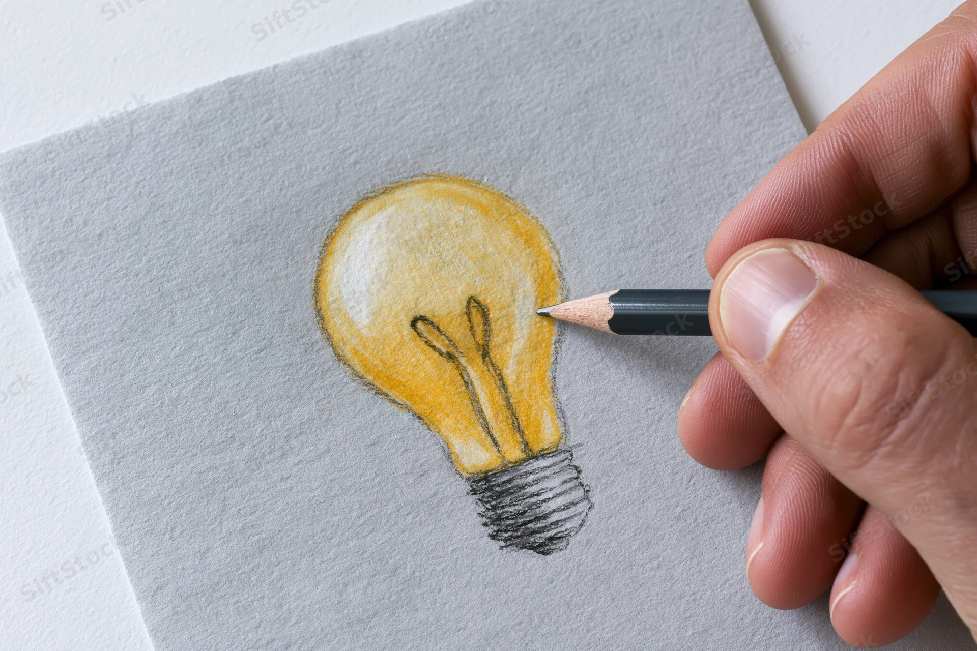 Hand drawing a glowing lightbulb on gray paper with a pencil