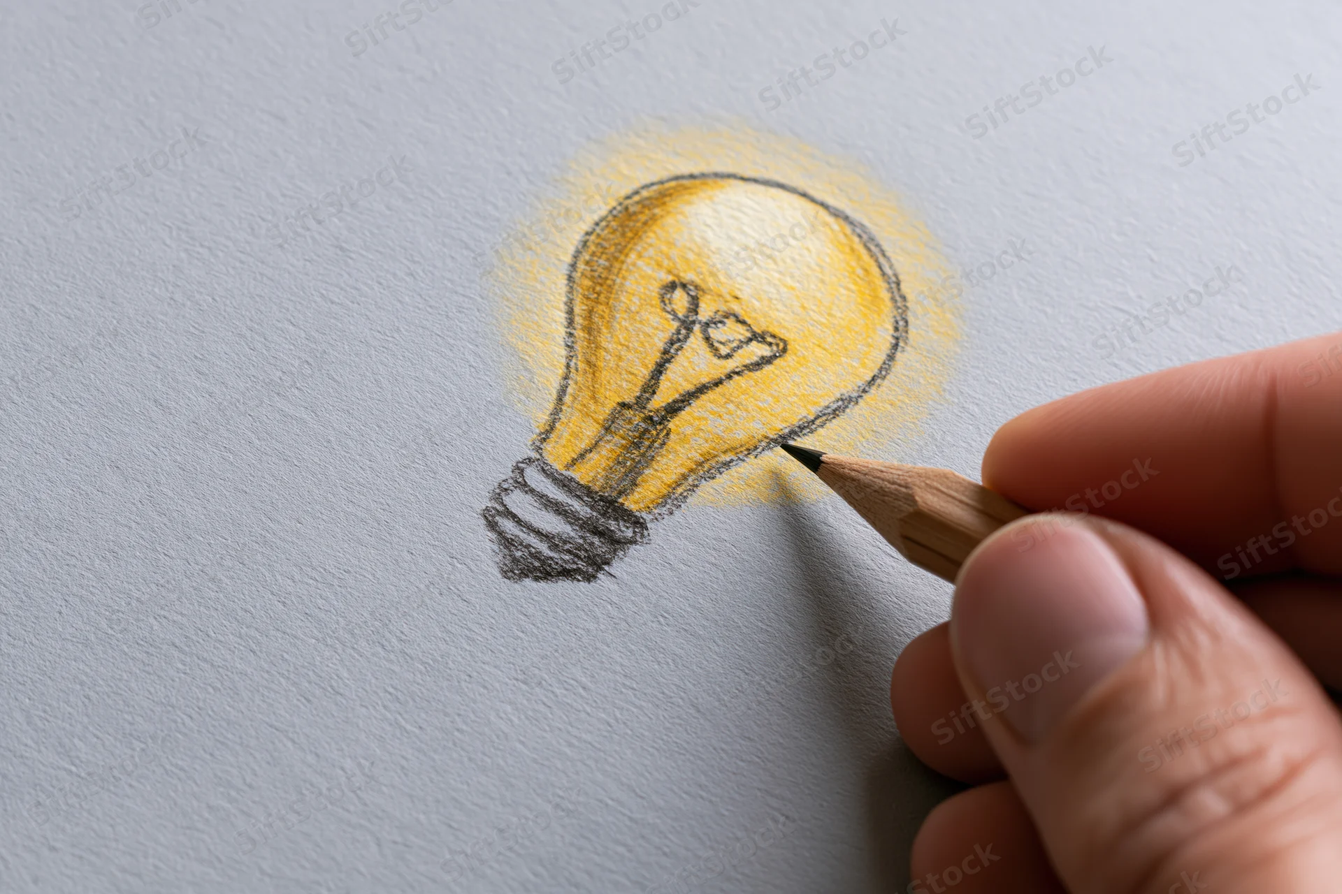 Hand drawing a glowing lightbulb on paper with a pencil