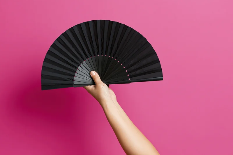 Hand holding open black folding fan raised upward against pink background