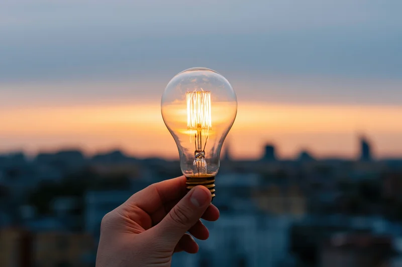 Hand holding lit filament lightbulb against city skyline at sunset