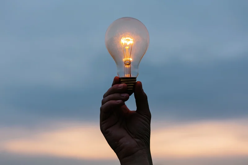 Hand holding glowing incandescent lightbulb against twilight sky